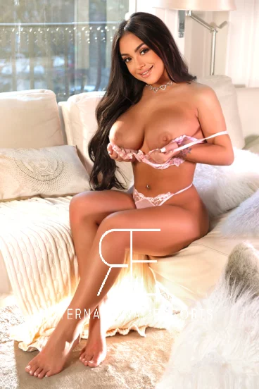 A very busty and sexy London escort is sitting on her sofa with her boobs out of her top 