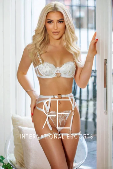 Blonde escort Alya is standing by a doorway and is dressed in white lace underwear 