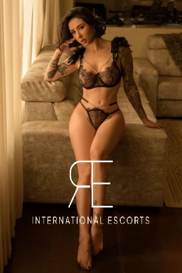Glamorous brunette in sheer black lingerie, seated with tattoos and irresistible allure.