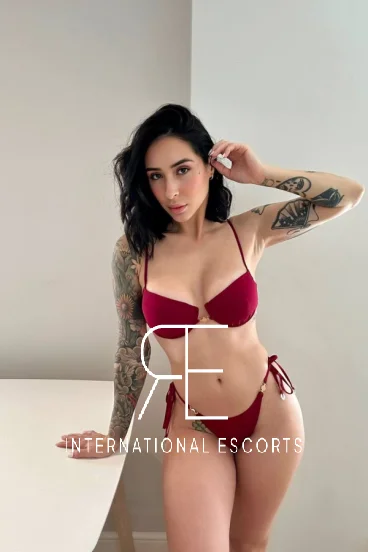 Tattooed escort Nya poses in a red bikini, her slim frame glowing in soft, natural morning light.