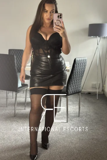 Lucy Rose in a black leather mini skirt, corset, and heels, confidently posing for a mirror shot.