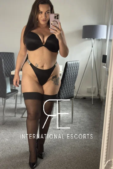 Lucy Rose in sleek black lingerie and heels, posing in a mirror selfie with a mischievous smile.