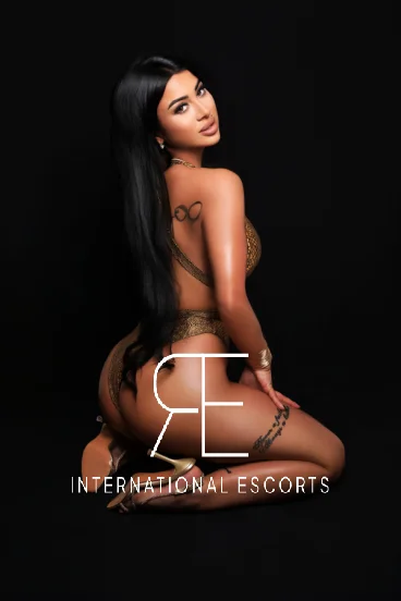 Brunette London escort in shimmering gold swimsuit on black backdrop